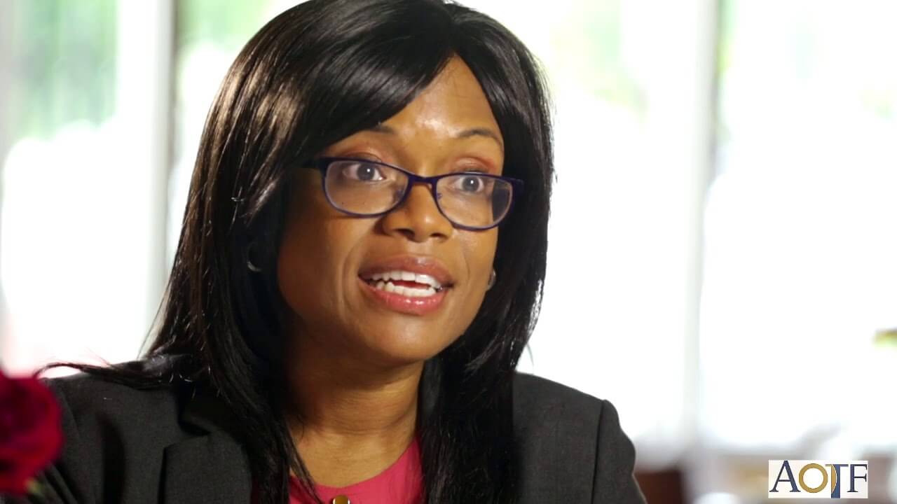 Professional woman wearing glasses and a black blazer speaking during an interview, seated indoors with soft natural light in the background.
