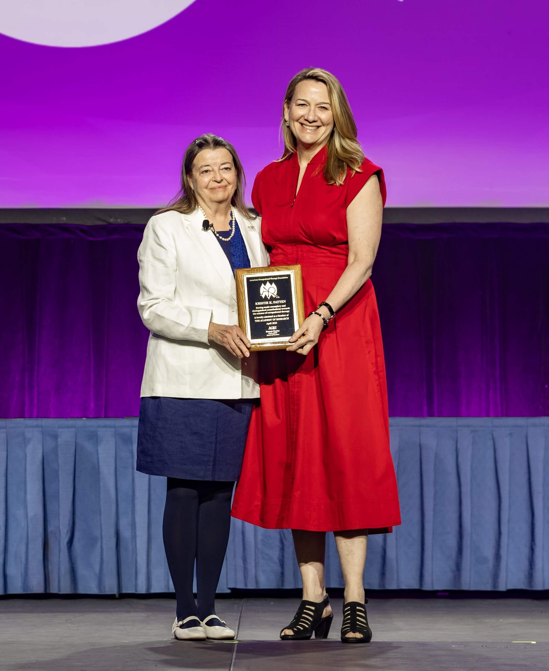A photo of our 2026 Academy of Research Nominee Kristie Patten being presented an award by our Board Chair Patti Davies on the AOTA conference stage with a purple backdrop in the background